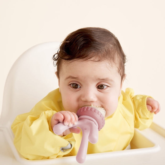 Eclectic B.Box blush silicone fresh food feeder held by a baby in a yellow bib sitting in a high chair