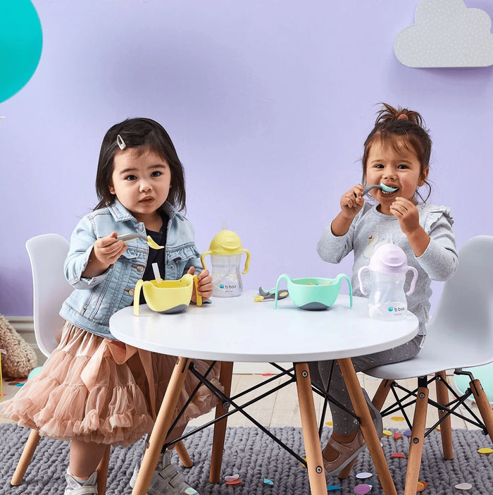 Eclectic b.box sippy cups and bowls on a table with two toddlers eating in a purple room