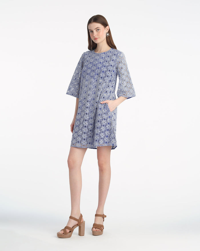 A woman stands against a white background wearing the Draper James Carolyn Bell Sleeve Mini Dress in Daisy Laceâ€”a blue and white, 1960s-inspired, knee-length dress with elbow-length sleeves and beige platform sandals.