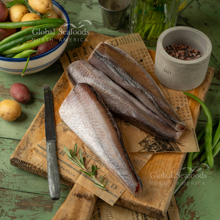 Headed and tailed Pacific whiting on a wooden board with herbs and vegetables nearby.