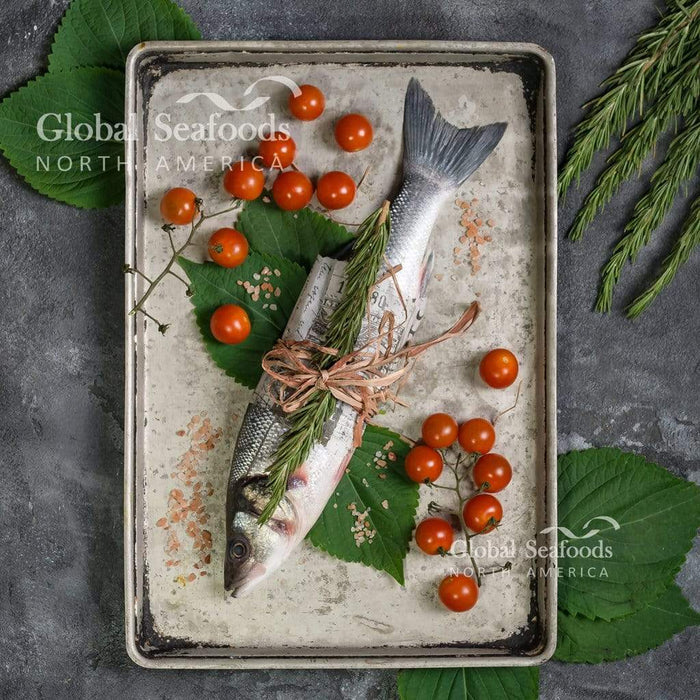 Whole branzino in a metal tray with tomatoes, herbs and green leaves.