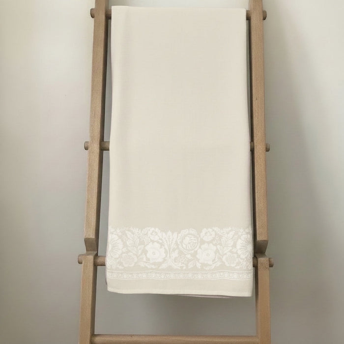 Eclectic natural beige linen-look tablecloth featuring an intricate white floral border draped on a wooden ladder.