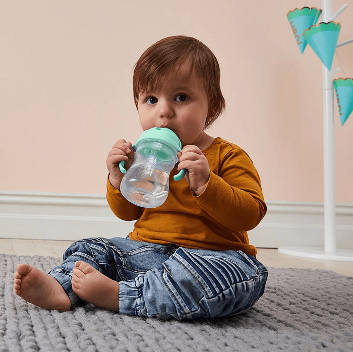 A toddler sitting on a rug holding a pistachio green b.box weighted straw sippy cup with easy-grip handles.