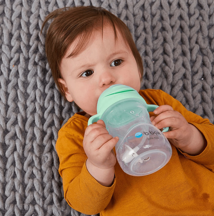 Eclectic b.box pistachio sippy cup with weighted straw held by a baby lying on a grey knitted blanket.