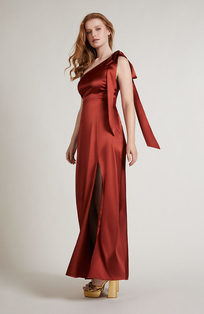 Woman wearing a burnt orange satin gown with a draped shoulder on a plain background