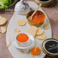Red trout caviar in a white bowl, with a crisp cracker topped with caviar in the foreground and an open caviar tin in the background, styled for a refined seafood snack presentation
