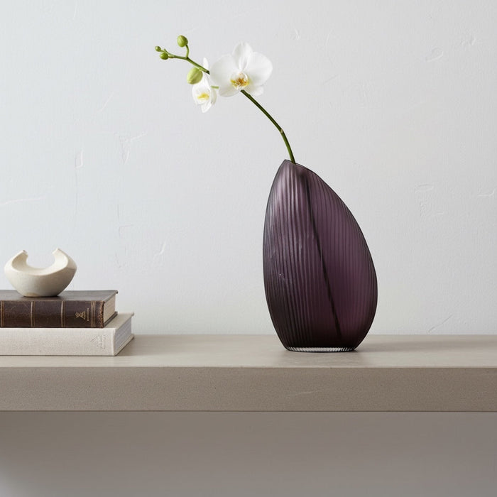 Eclectic violet ribbed glass vase with an asymmetrical silhouette holding a white orchid on a shelf.