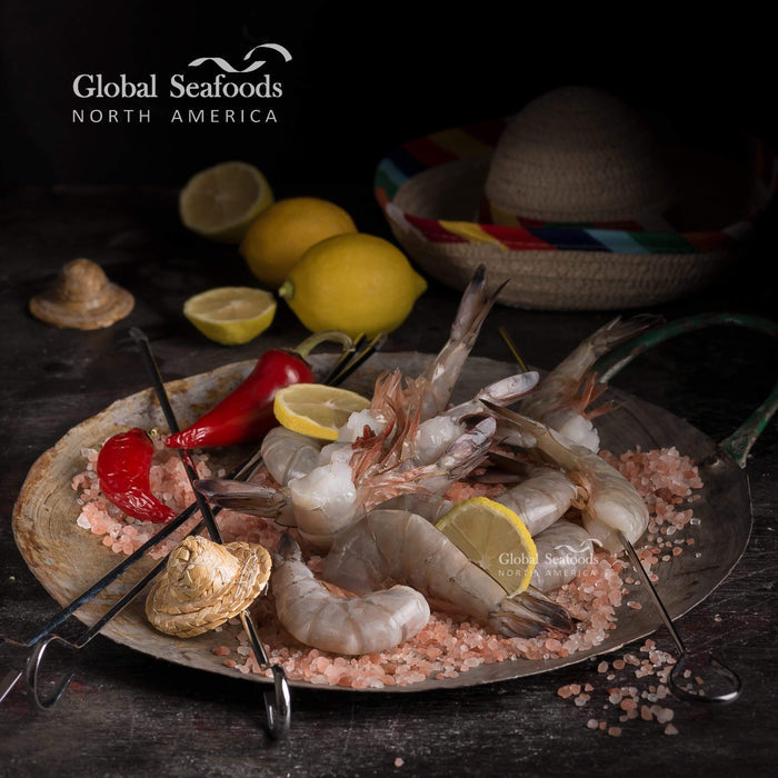 Fresh Wild Blue Mexican shrimp tails arranged on a dark metal plate with pink salt, skewered on metal BBQ sticks and garnished with lemon slices. Whole lemons and a traditional Mexican hat sit in the background, all set on a dark table for a bold and festive seafood presentation