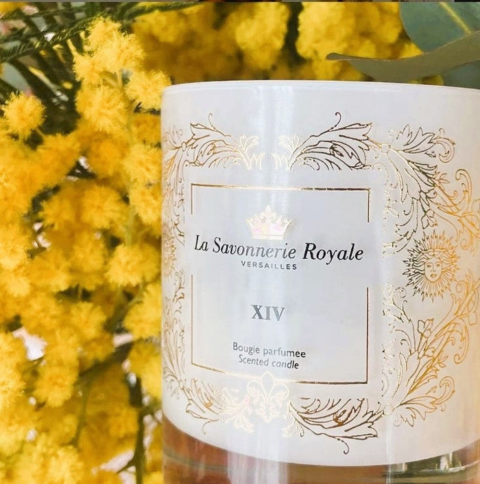 Eclectic La Savonnerie Royale XIV scented candle in a white glass vessel with ornate gold detailing near yellow flowers.